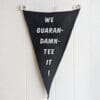 black pennant with two metal grommets embroidered in white with the phrase "we guaran-damn-tee it"