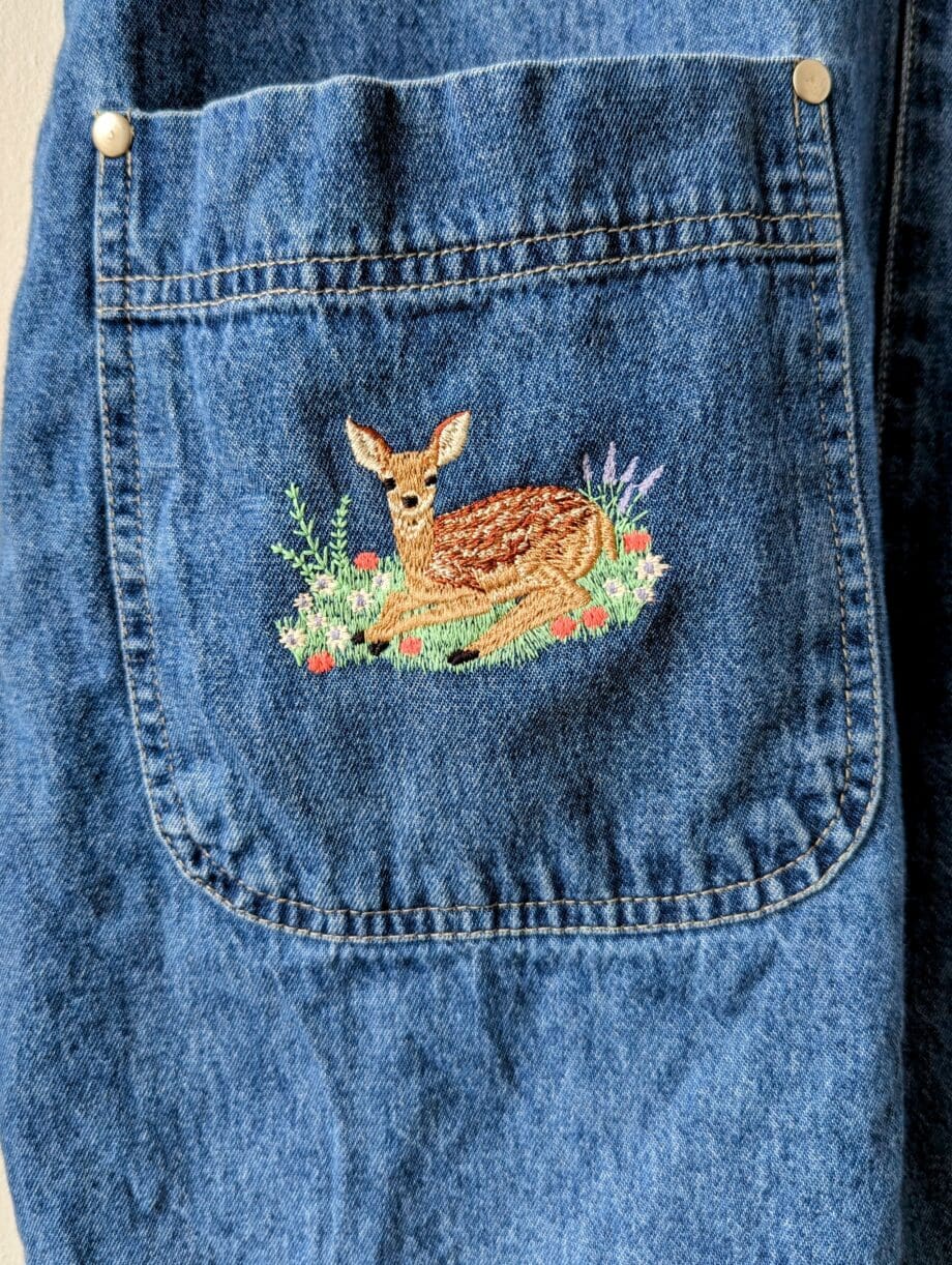 Detail of deer embroidery on front right pocket. Deer is in a bed of grass and flowers and lilac.