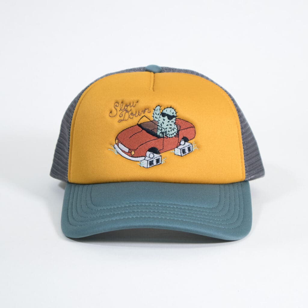 trucker hat with a mesh back in forest green with a mustard yellow front panel embroidered with a cactus wearing sunglasses and waving in a car that is sitting on cinderblocks instead of wheels with the slogan "Slow Down."